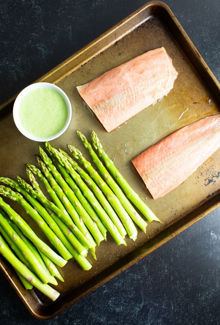 Raw salmon and asparagus with a small bowl of cilantro lime sauce arranged on a sheet pan.