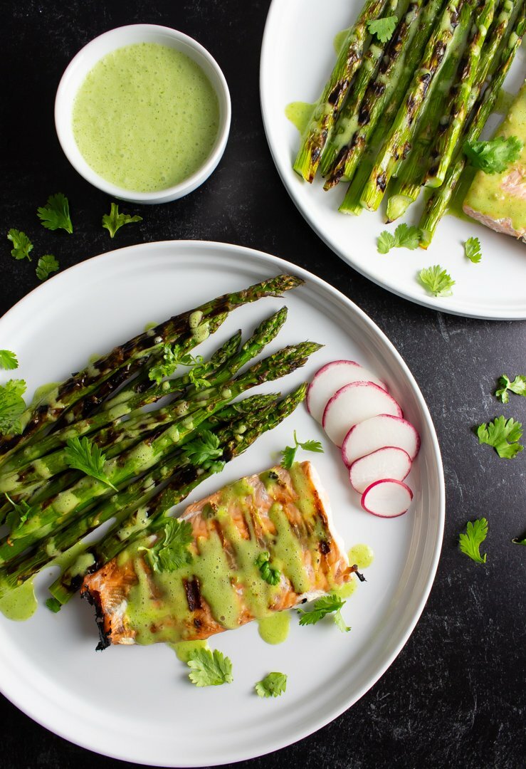 Top view of Grilled Salmon and Asparagus with a small bowl of Cilantro-Lime Sauce on white plates with a textured black background.