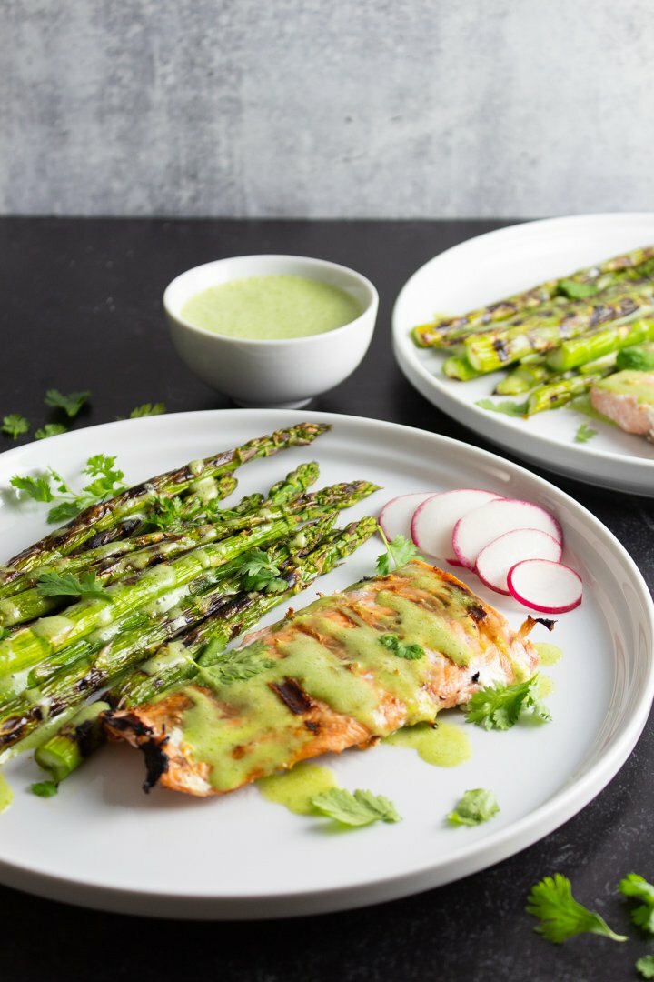 Side view of Grilled Salmon and Asparagus with a small bowl of Cilantro-Lime Sauce on white plates with a textured black background.